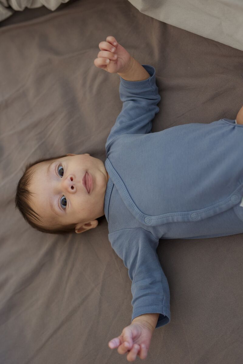Baby in blue onesie looking up from the bed